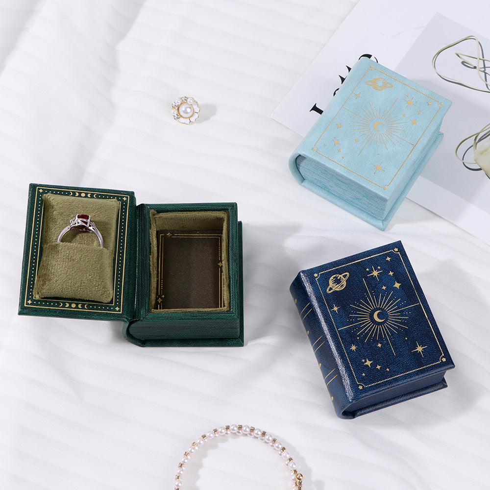 Open jewelry box with a ring, closed blue book-shaped box, and bracelet on a white surface.